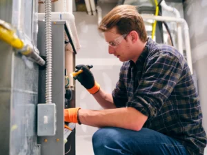  A man wearing a plaid shirt and safety glasses is diligently working on a furnace, ensuring safety and precision.